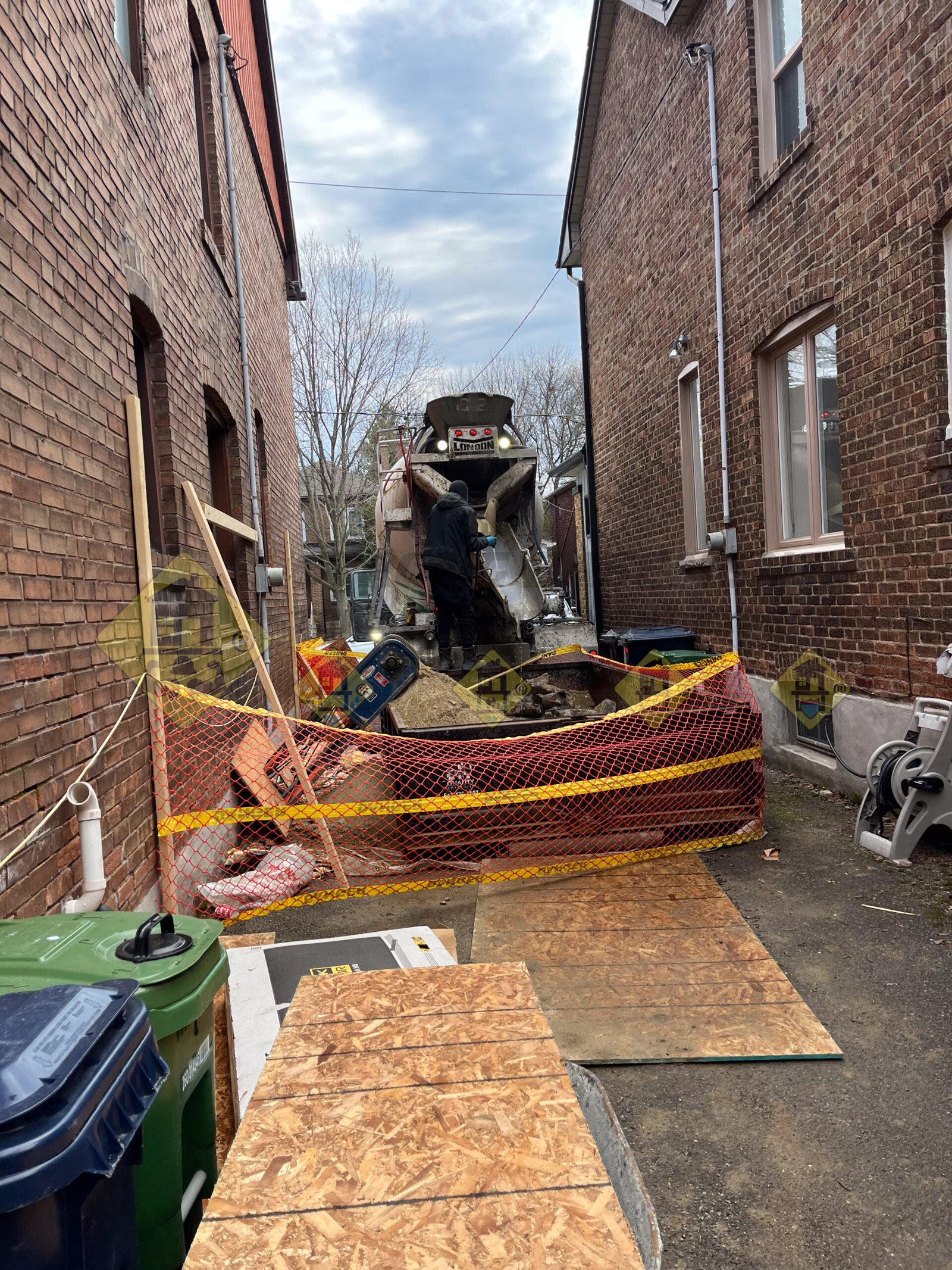 A concrete truck parked in front of a full 6-yard soil disposal bin in a narrow Leaside laneway, alongside a safely fenced conveyor belt setup used for a basement underpinning project.