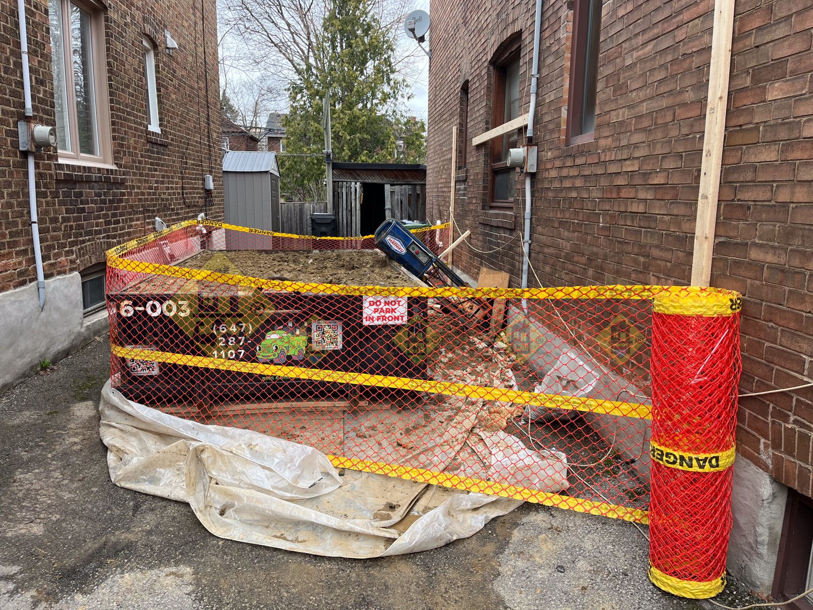 A 6-yard soil disposal bin full of dirt in a narrow Leaside laneway, fed by a conveyor belt from a side window, and surrounded by safety fencing during a basement underpinning project.